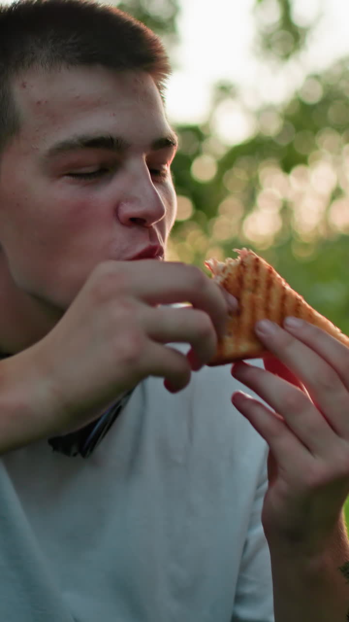 hombre con mano tatuada haciendo un brindis con pan, con una vista parcial de otra persona sosteniendo pan en el fondo, la luz del sol se filtra a través de los árboles, creando una cálida atmósfera al aire libre