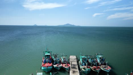 jetty looking out towards an island in the sea with moored colourful fishing boats, drone aerial fly over