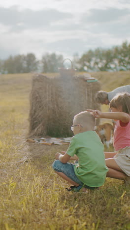 una mujer se agacha para tomar una foto de los niños jugando mientras un amigo extiende una manta mientras un perro juguetón se sube a ella, el entorno rural iluminado por el sol captura el vínculo familiar alegre y la diversión al aire libre