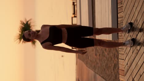 vertical video: a brunette girl with tied hair in a black sports uniform does squats on a sunny beach with palm trees, a beach covered with boards