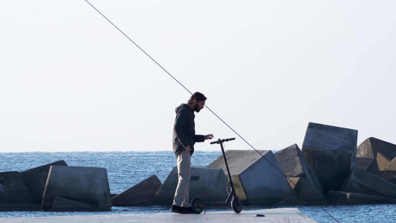 el hombre se baja del scooter eléctrico para tomar una llamada telefónica por el océano, se dirige hacia la cámara