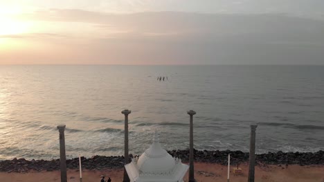 Toma-Aérea-De-La-Estatua-De-Mahatma-Gandhi-Desde-La-Playa-En-Un-Amanecer-Dorado-En-La-Mañana-Cerca-De-La-Playa-De-Pondicherry-Rock,-Filmada-Con-Un-Dron-En-4k