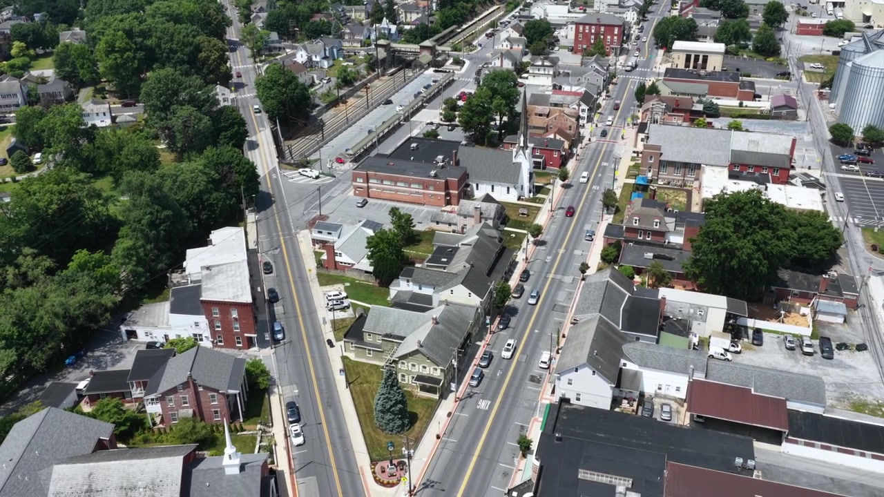 Incrocio stradale a Y divide i tetti fitti di una piccola città a Mount Joy, Pennsylvania; auto percorrono strade divergenti oltre un edificio scolastico in mattoni e alberi verdi e ombrosi. Ripresa aerea con drone all'indietro.