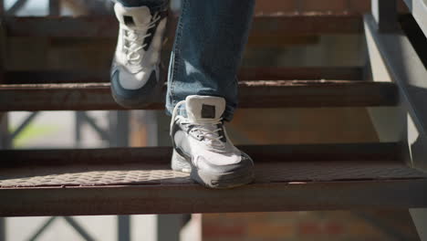 close up of jeans clad legs descending metal staircase wearing black white canvas sneakers with white socks stepping down rusted steps against brick and metal railing in sunlit urban scene