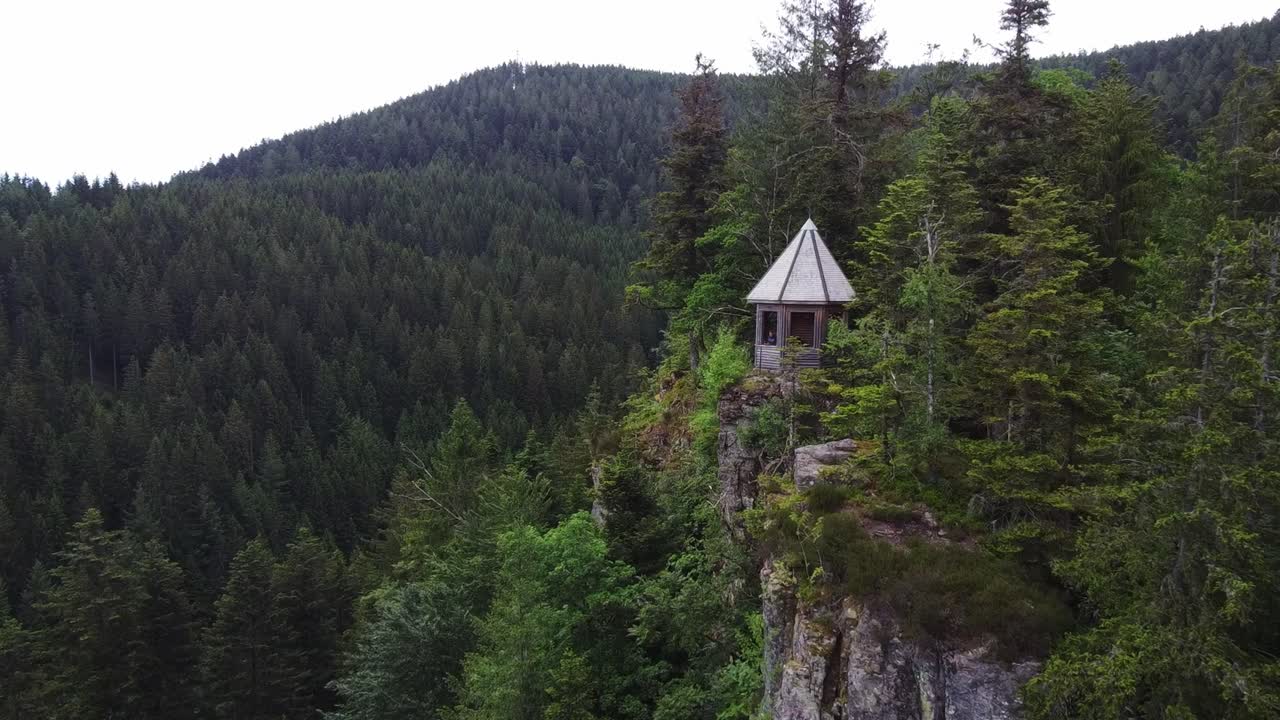 An aerial view captures a gazebo nestled on a cliff at the top of a mountain, surrounded by a dense forest. The view conveys a sense of tranquility and natural beauty.