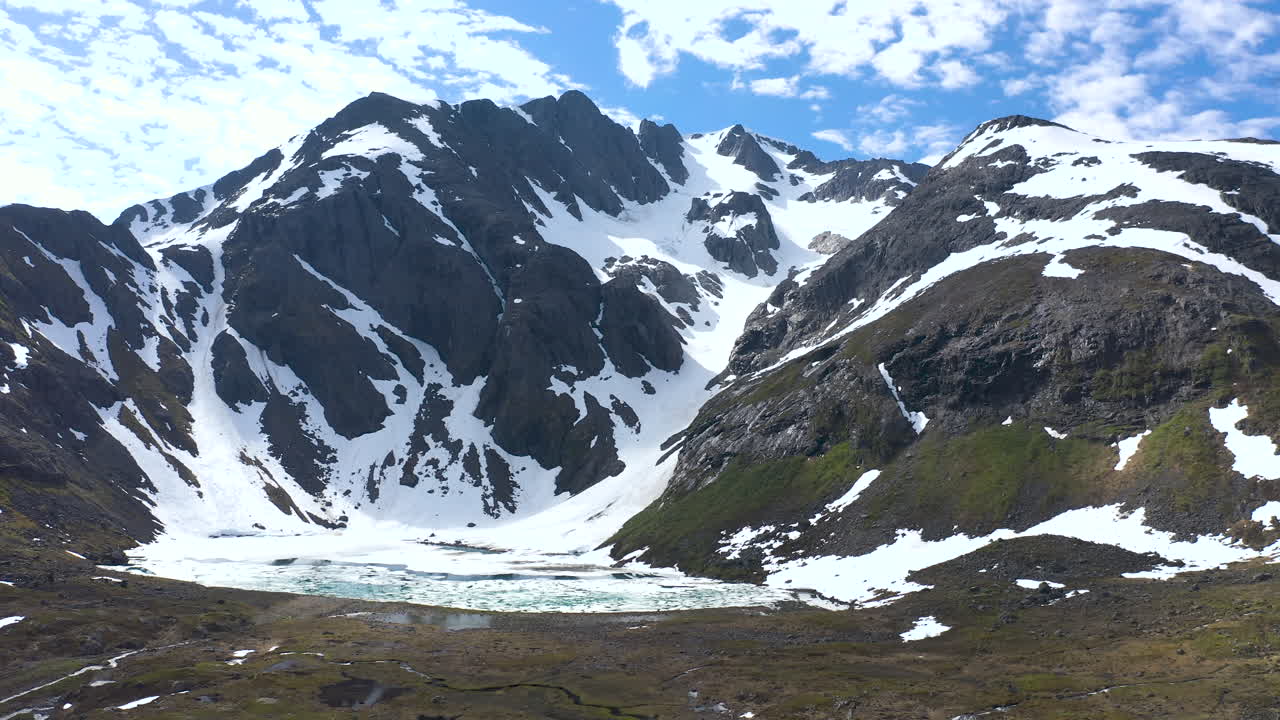 Aerial shot approaching slowly towards a mountain where snow and glacier ice has almost completely melted as background show blue sky with clouds