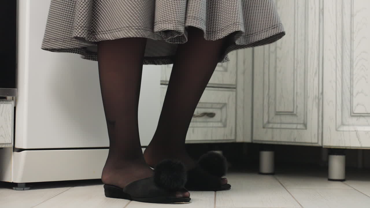 leg view of lady in black pantyhose and designer slippers with floating gown dancing gracefully near kitchen cabinet, expressing elegance and style with smooth movement in modern home