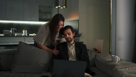 A-beautiful-brunette-girl-in-a-beige-T-shirt-is-interested-in-how-his-husbands-work-is-going-remotely.-A-brunette-with-stubble-in-a-black-jacket-and-beige-shorts-sits-on-a-gray-sofa-in-a-modern-studio-apartment-in-the-evening