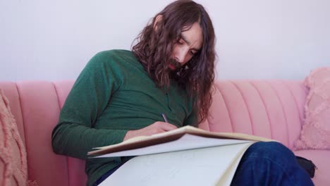plano medio en cámara lenta del artista masculino hispano con cabello largo y barba sentado en un sofá rosa dibujando y borrando en un bloc de bocetos