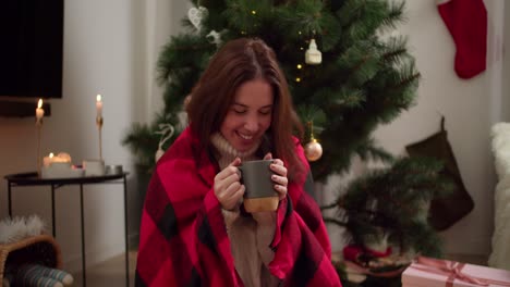 Happy-brunette-girl-in-a-red-checkered-plaid-drinks-hot-tea-from-a-gray-mug,-smiles-and-looks-to-the-side-near-the-Christmas-Tree-in-a-cozy-room-in-the-evening-in-winter