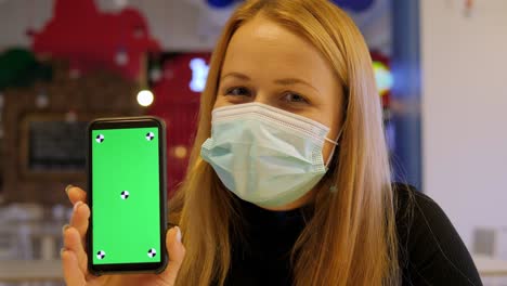 a woman in a medical mask holds a phone with a chroma key in a cafe.