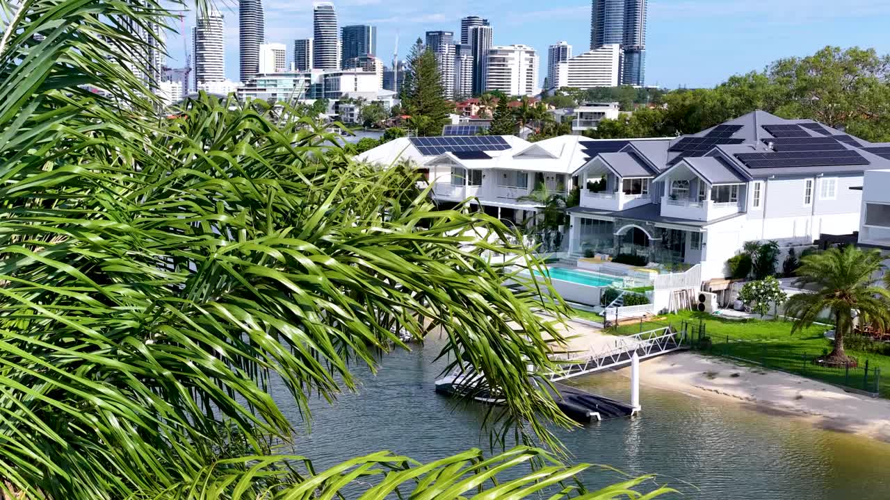 Camera pans past palm leaves to reveal canal, upscale homes, boats, and Gold Coast skyline