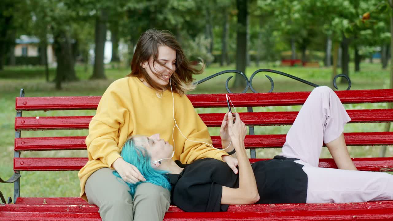 Veninder lytter til musik på en bænk. Lesbisk par taler og lytter til musik, mens de slapper af på en bænk en sommerdag i parken.