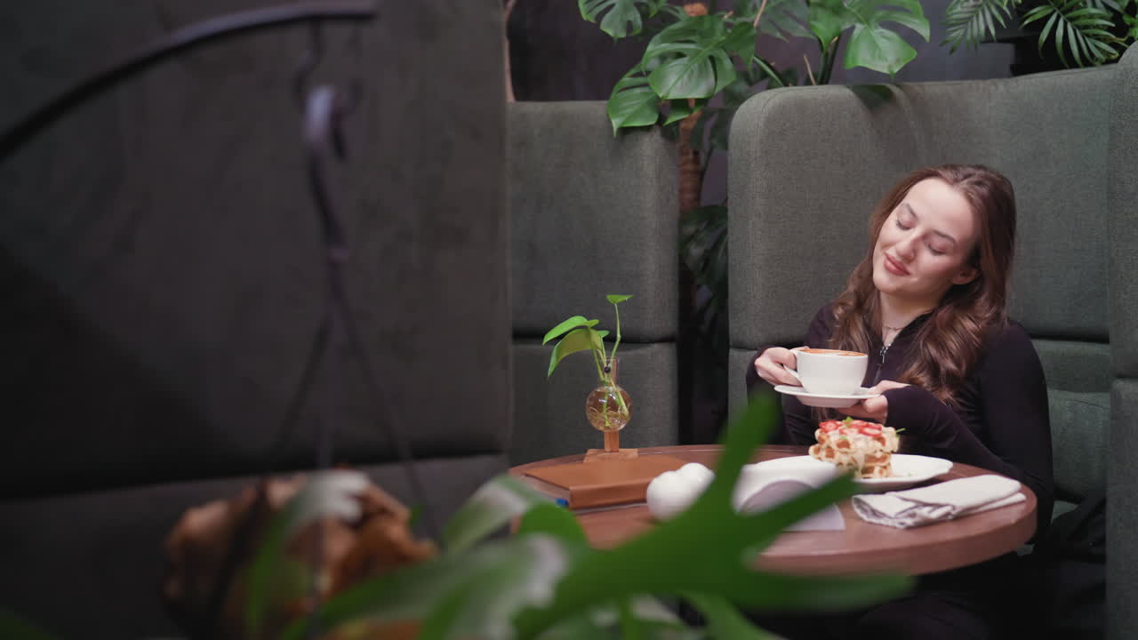 Young woman in cozy booth holds white cup of latte close to face with gentle smile, eyes closed while enjoying aroma, surrounded by indoor plants, waffles on plate, visible in background