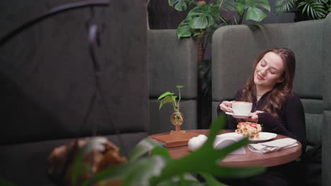 young woman in cozy booth holds white cup of latte close to face with gentle smile, eyes closed while enjoying aroma, surrounded by indoor plants, waffles on plate, visible in background