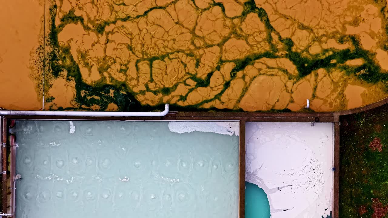 Tampilan atas ke bawah mengontraskan kolam alga oranye cerah dengan tangki pengolahan air berwarna pirus, yang dipisahkan oleh struktur pipa putih mencolok