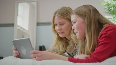 mutter mit teenager-tochter in pyjamas, die auf dem bett liegt, überträgt film oder video auf ein digitales tablet - in slowmotion gedreht