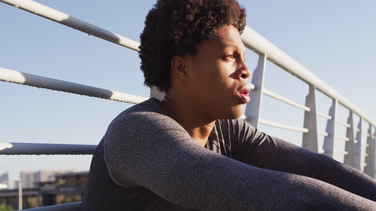 Fit african american man exercising outdoors in city, resting sitting on footbridge