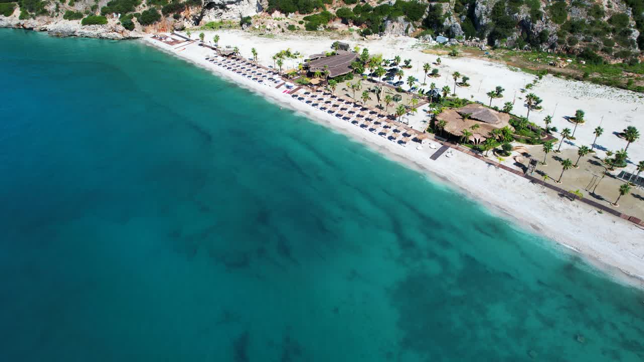 Beach bar restaurants on shore with white pebbles and palms garden resort, turquoise Ionian sea, Albania