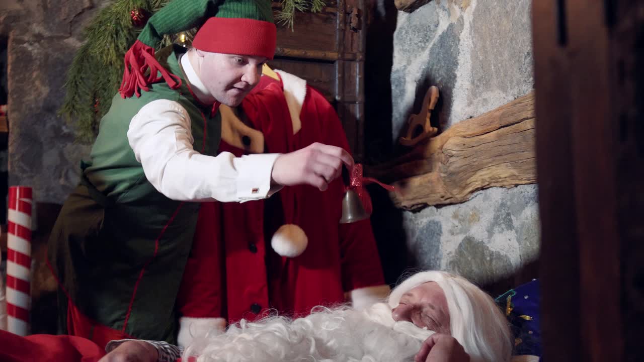 Santa with white beard sleeping in his residence. Funny elves ringing with a little bell to wake up Santa. Christmas time