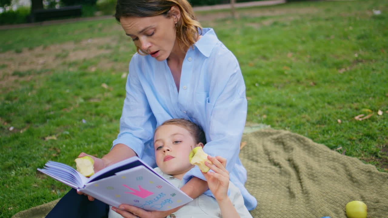 Picnic family reading book on green grass at weekend closeup. Kid eating apple