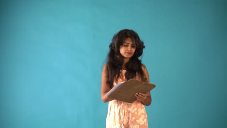 una joven india con vestido naranja pensando con un bloc de notas y un lápiz de pie en un fondo azul aislado