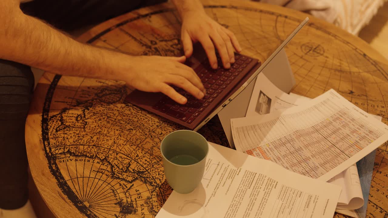 A man sits at a wooden table and starts working on his laptop surrounded by papers and a coffee cup. Cozy home office atmosphere in warm lighting