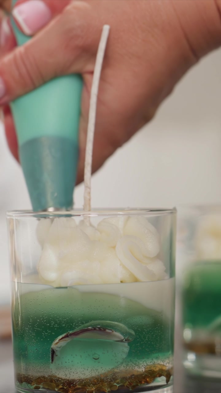 Primer plano de una mano usando una manga pastelera aguamarina para llenar portavelas de vidrio con cera blanca sobre líquido verde. La escena presenta múltiples velas en varias etapas, colocadas sobre una superficie lisa bajo una luz suave.