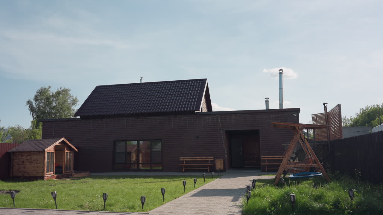Brown wooden house in sunlit fenced compound with wooden cabin, green lawn swaying in breeze, paved walkway to bench seating and wooden swing under clear sky with light clouds