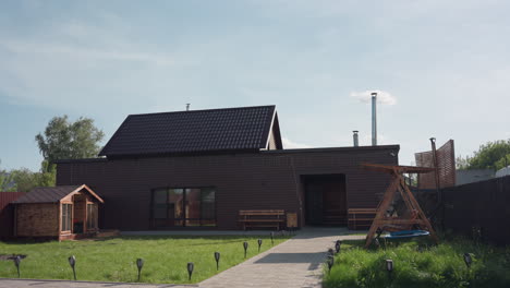 brown wooden house in sunlit fenced compound with wooden cabin, green lawn swaying in breeze, paved walkway to bench seating and wooden swing under clear sky with light clouds