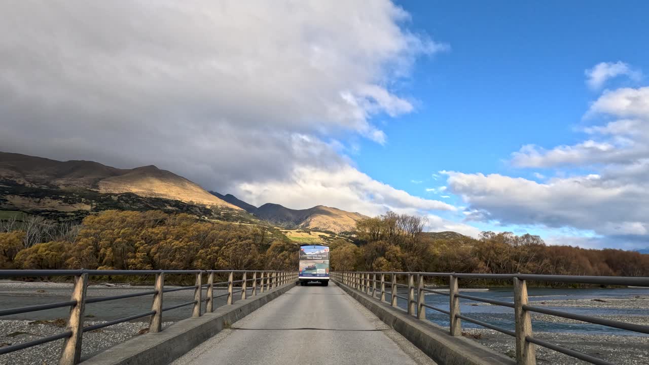 Campervan drives across rural bridge with mountains, river, and dramatic clouds in natural daylight