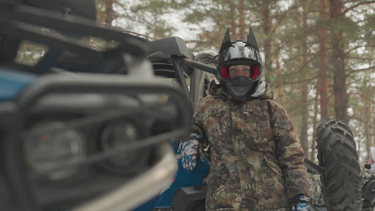 Pilota caucasico accanto alla parte anteriore dell'ATV, faro affilato e griglia blu in primo piano, pilota con casco sfocato e mimetica nella foresta di pini, ispezione del veicolo e preparazione meccanica prima dell'off-road estremo.