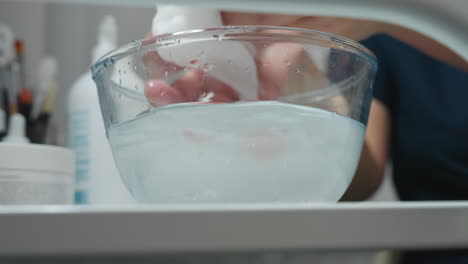 hand view of facialist dipping cotton pad into glass bowl of purified water on white trolley beside skincare bottles, then gently pressing water from pad over facial bed in clinic room