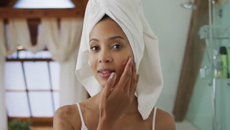 Portrait-of-mixed-race-woman-wearing-towel-on-head-applying-cream-on-her-face