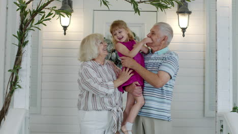 Happy-senior-grandfather-and-grandmother-couple-holding-granddaughter-in-hands-in-porch-at-home