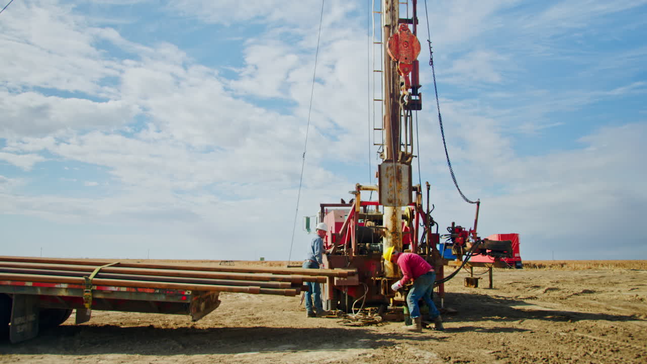 Two men work near the drilling equipment in the valley. Workers attach the heavy metal detail with the help of crane.