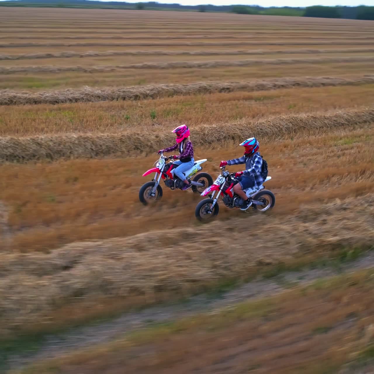 Young couple riding motorcycles close to each other. People having fun in the field with mowed grass at setting sun. View from above