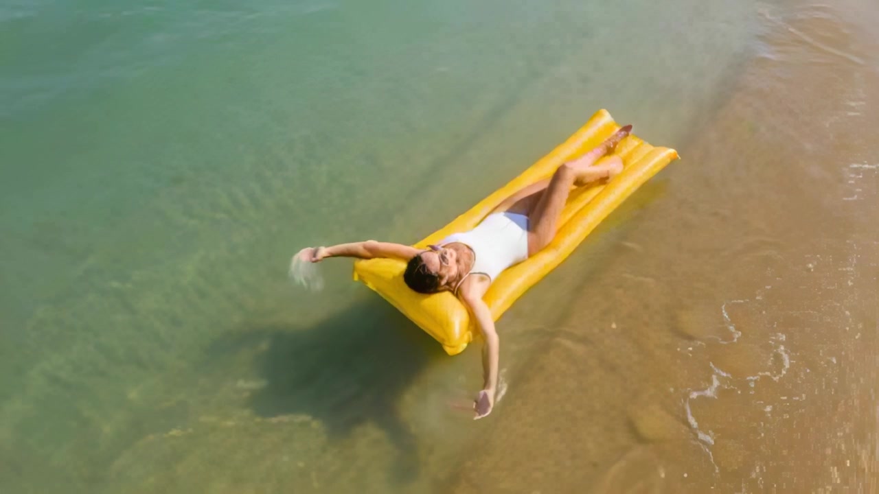 Relaxing woman in white-one-piece swimsuit skimming water on yellow mattress at beach as waves lap