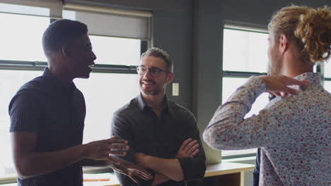 Four-happy-male-creatives-stand-talking-in-their-workplace,-waist-up,-close-up