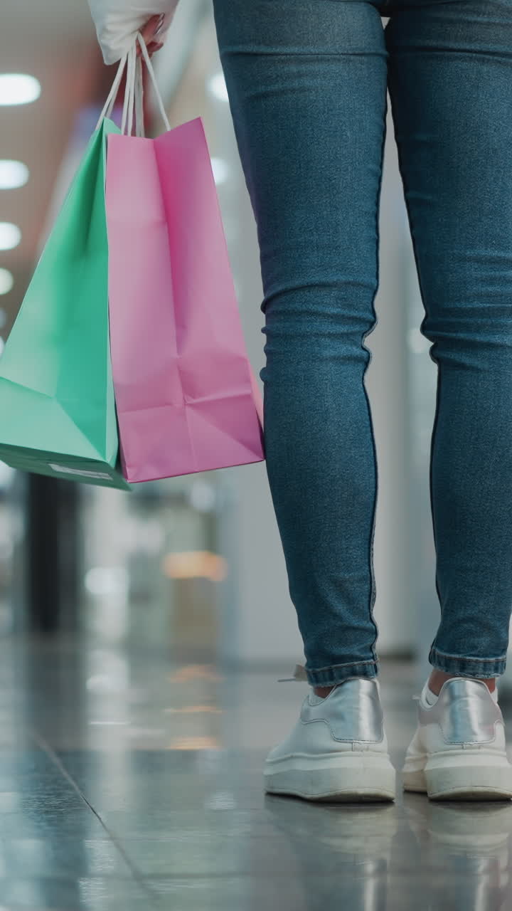 잘 조명 된 쇼핑몰에 서 있는 여성이 들고 있는 쇼핑 가방의 클로즈업, 한 가방은 분홍색이고 다른 가방은 초록색이며, 그녀는 흐릿한 배경으로 반이는 바닥을 걸어가면서 부드럽게 흔들립니다.