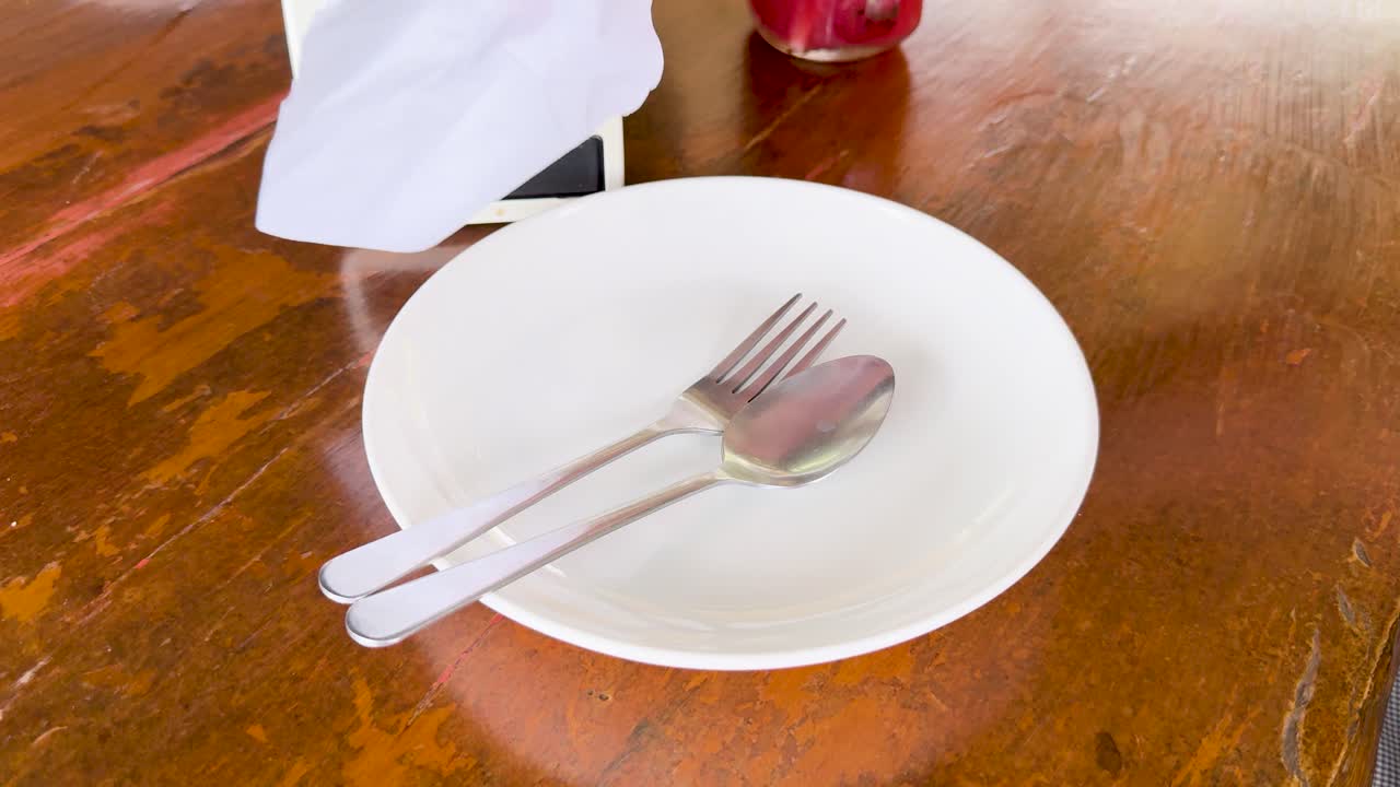 Hands arrange cutlery and napkin on a plate at a wooden table in natural light