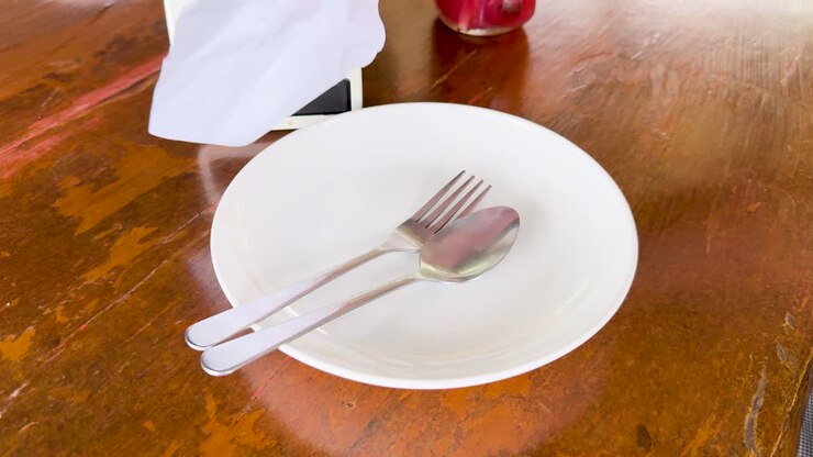 Hands arrange cutlery and napkin on a plate at a wooden table in natural light