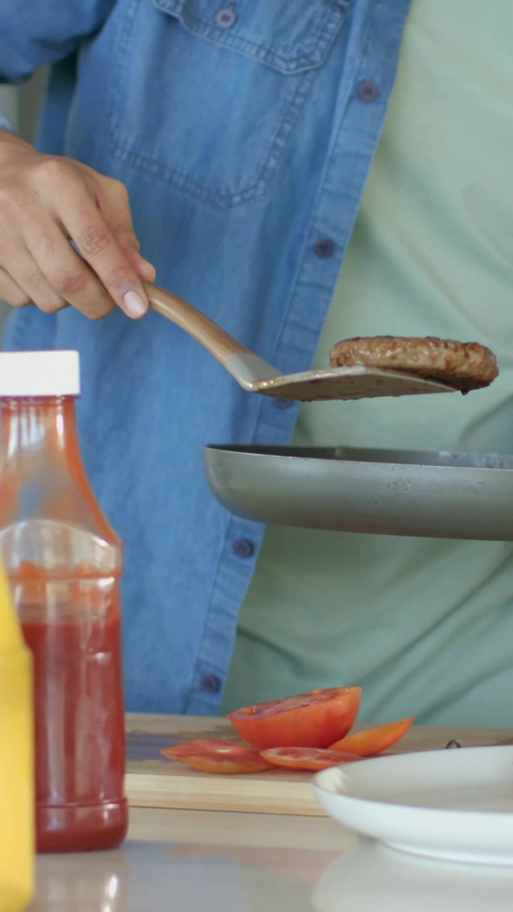 Video verticale: Coppia in denim e rosa che solleva un hamburger dalla padella, assemblando un panino a casa per il pasto.