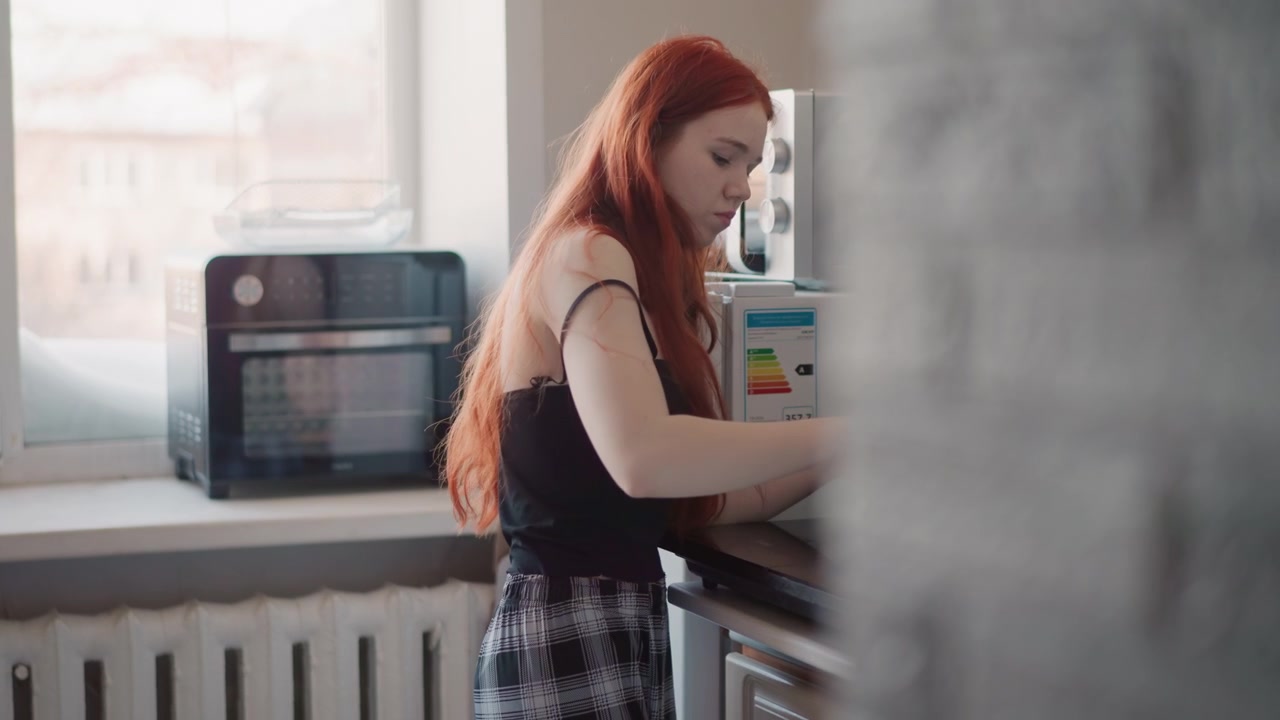 Retrato de mujer caucásica en cocina iluminada por el sol, inclinada sobre el fregadero, cabello rojo largo cerca de un horno tostador, luz natural suave, expresión contemplativa, ritual matutino privado que insinúa previsión y una fortuna tranquila