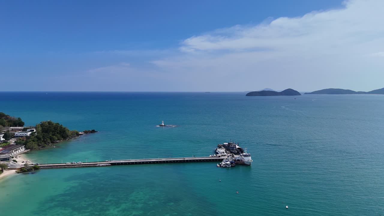Drone footage captures a serene Phuket coastline with clear blue waters, a pier, and distant islands under bright daylight
