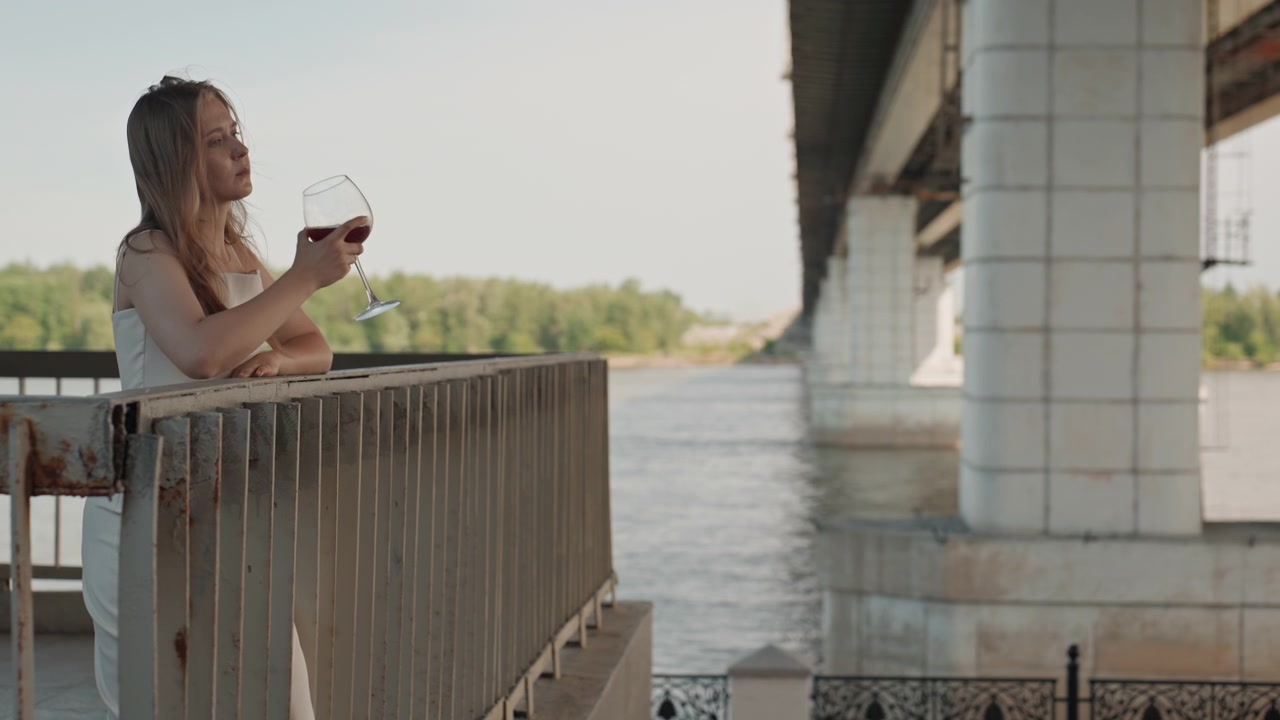 Donna in abito bianco sta su un ponte, tenendo in mano e sorseggiando vino rosso, mentre ammira l'acqua e la vegetazione. La scena presenta una luce naturale soffusa e un'espressione calma, con il soggetto ben posizionato.