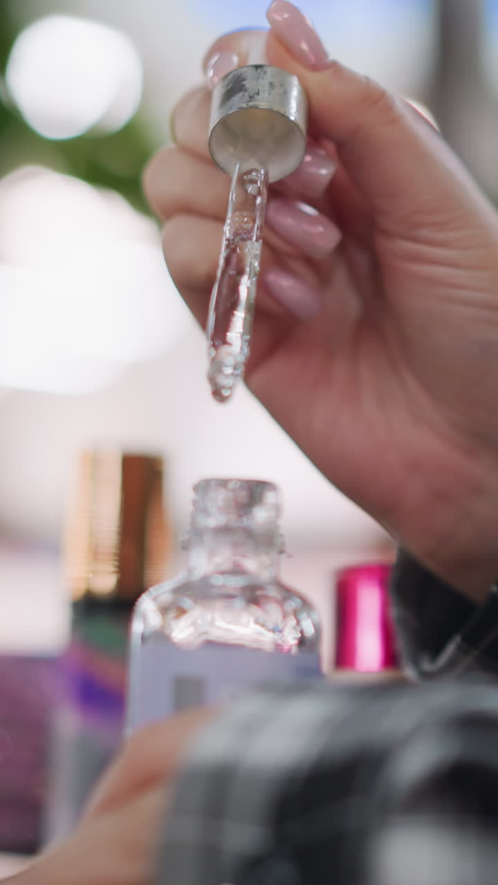 Closeup precise serum dropper pouring into bottle, elegant hand with glossy manicure holding silver cap while clear viscous serum forms steady droplet, sleek packaging and metallic accents, studio
