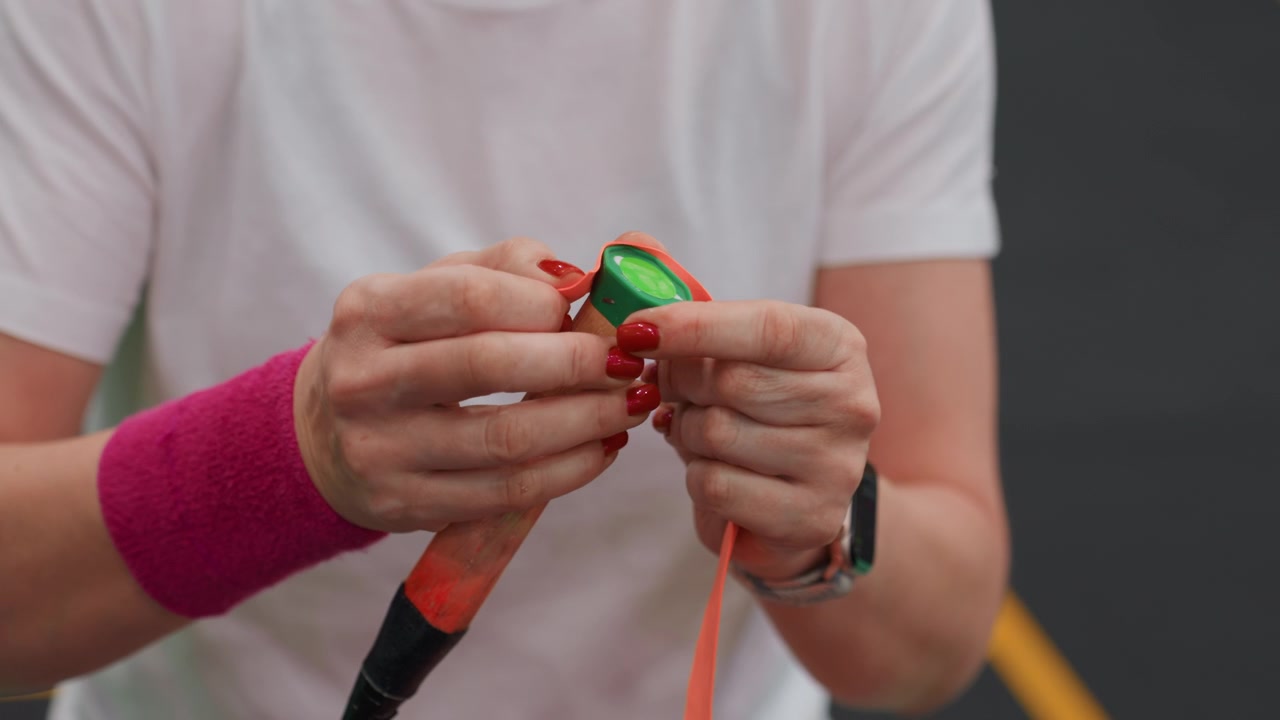 Primo piano di mani che applicano un antivibrazione da badminton, donna bianca che monta un antivibrazione sul manico della racchetta, polsino rosa, smartwatch, campo al coperto, preparazione meticolosa per l'allenamento e dimostrazione di coaching.