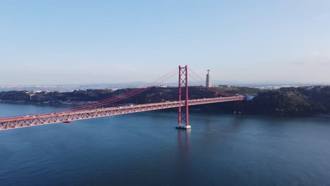 vuelo de un dron por encima del agua en portugal con puente colgante rojo y monumento de cristo en segundo plano