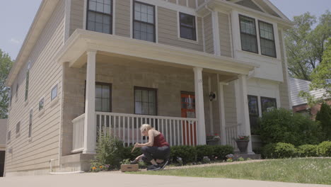 a wide low angle shot of a woman planting flowers in a flower bed in front of her house